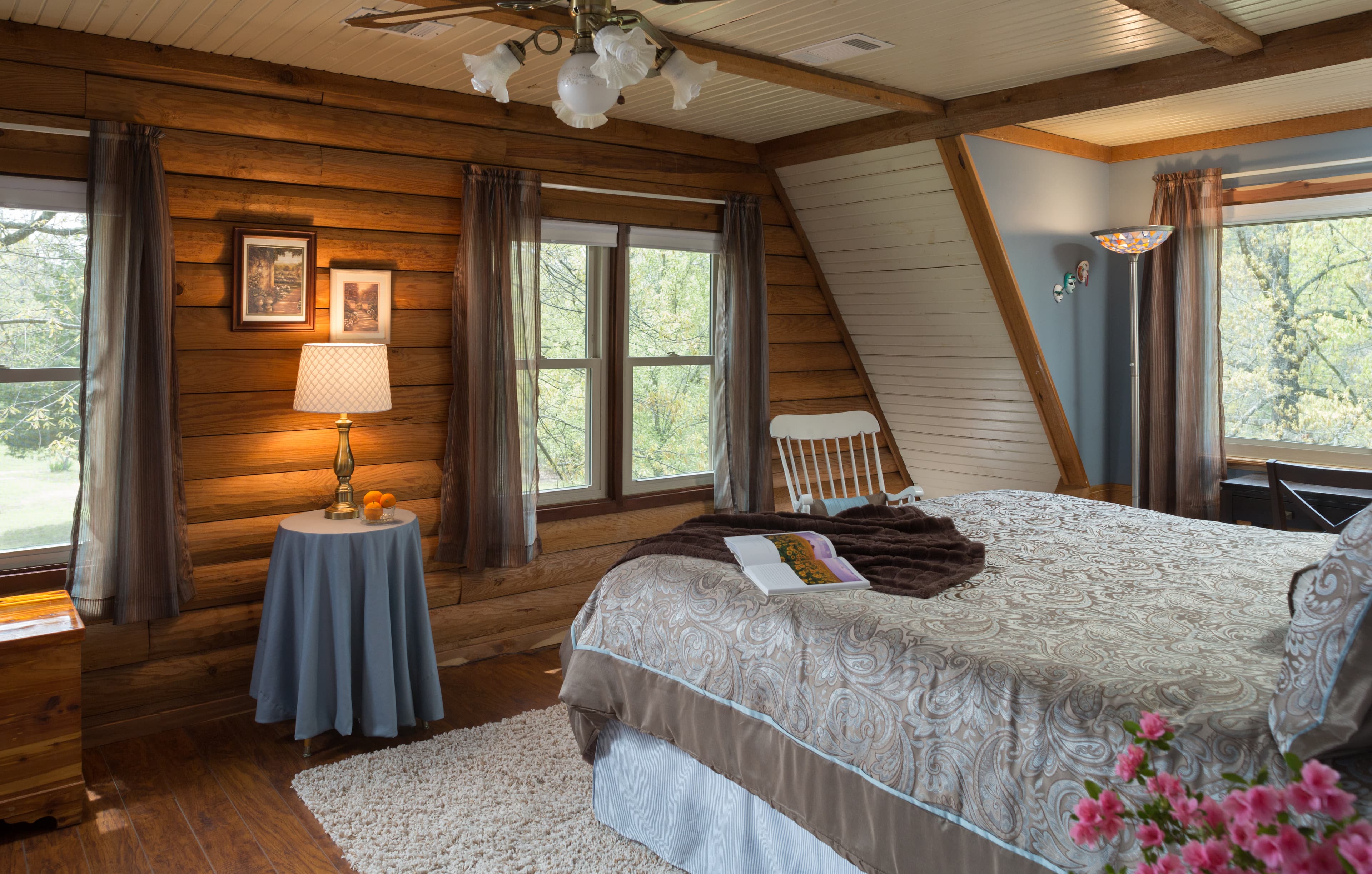 king size bed in front of a large picture window with green trees on the outside.  Facing wall consists of log, with 2 windows.  In between the windows is a small round table with a blue table cloth and a brass table lamp.  White rocking chair in corner