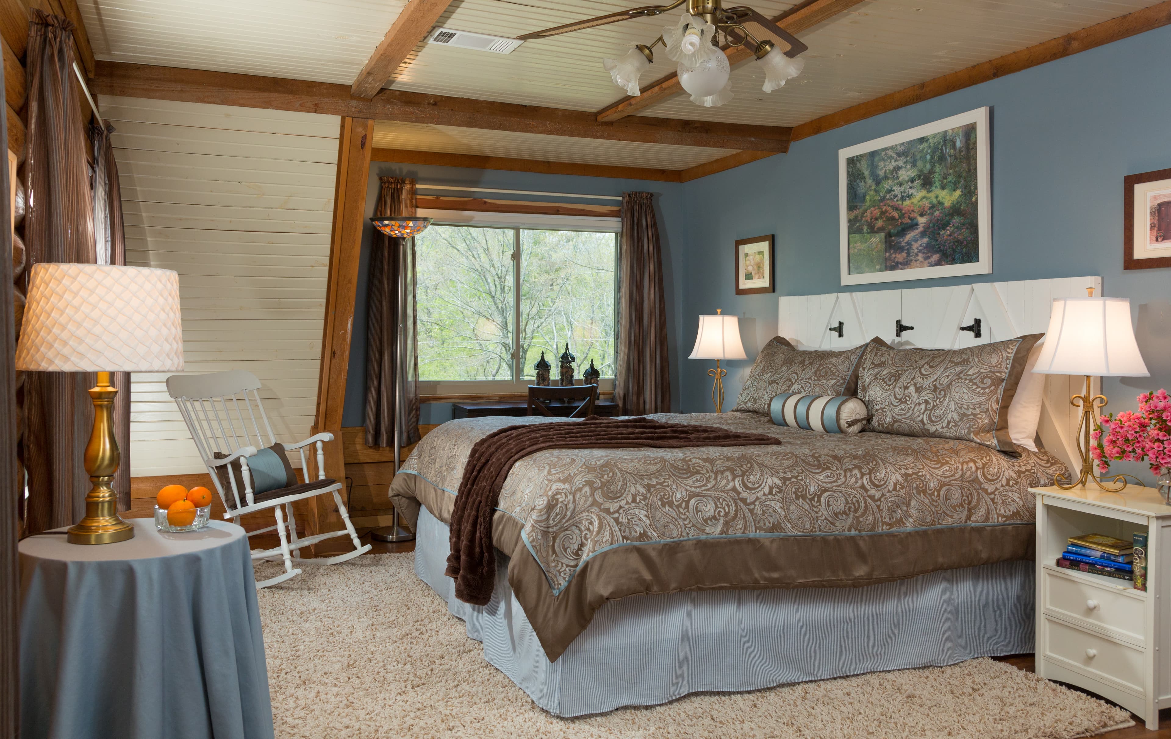 king-sized bed with 2 large and one small accent pillows, a brown throw blanket, and a brown & blue duvet cover.  To the right of the bed is a white nightstand wtih books on the shelf, and a table lamp and pink flowers on top.  To the left of the bed is a matching lamp, and a small desk in front of a large window.