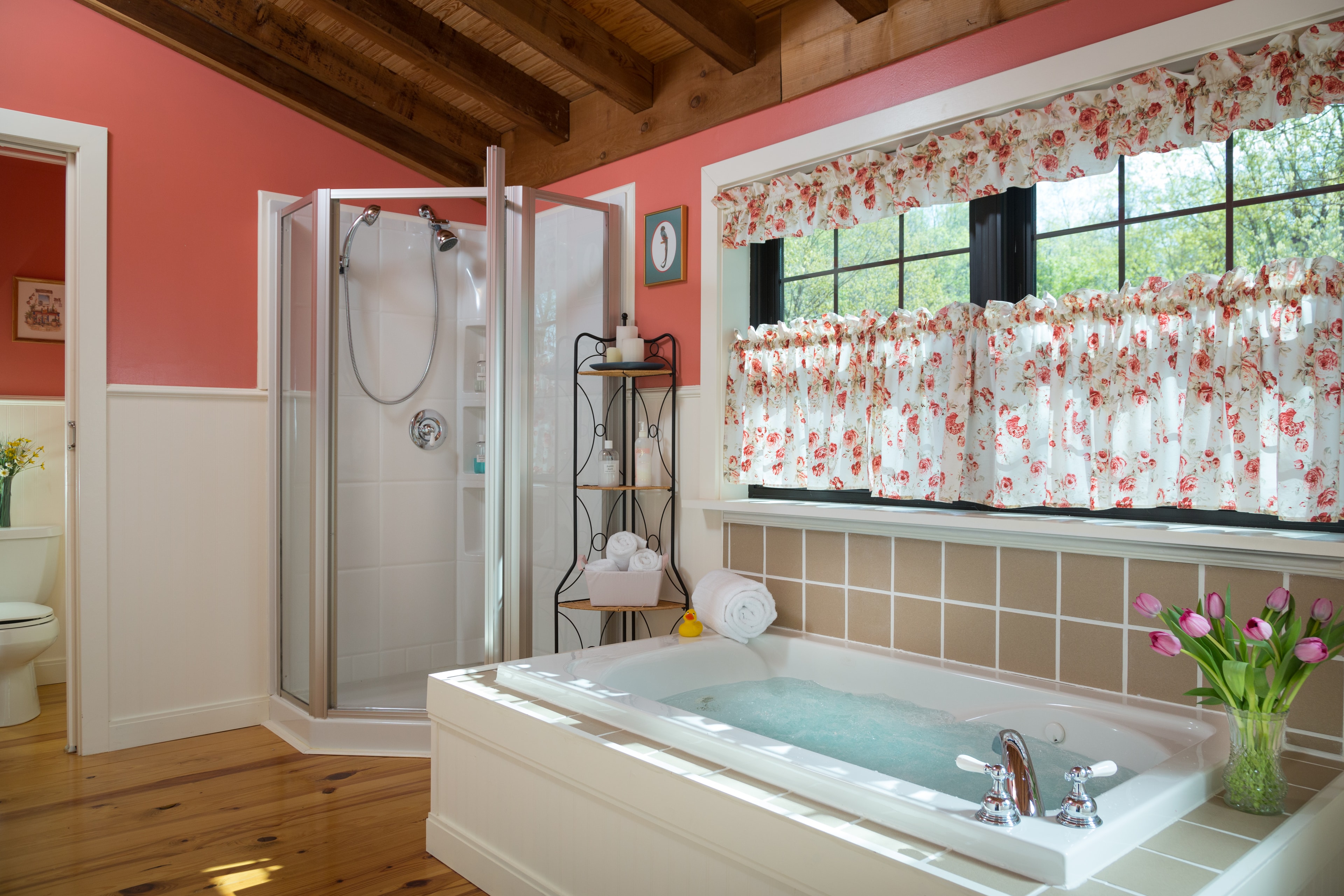 Large bathroom with a stand alone shower with a glass door. Next to the shower is a small corner shelf with bath accessories. To the right of the shelf is a jetted tub filled with water, and windows with pink & white curtains.