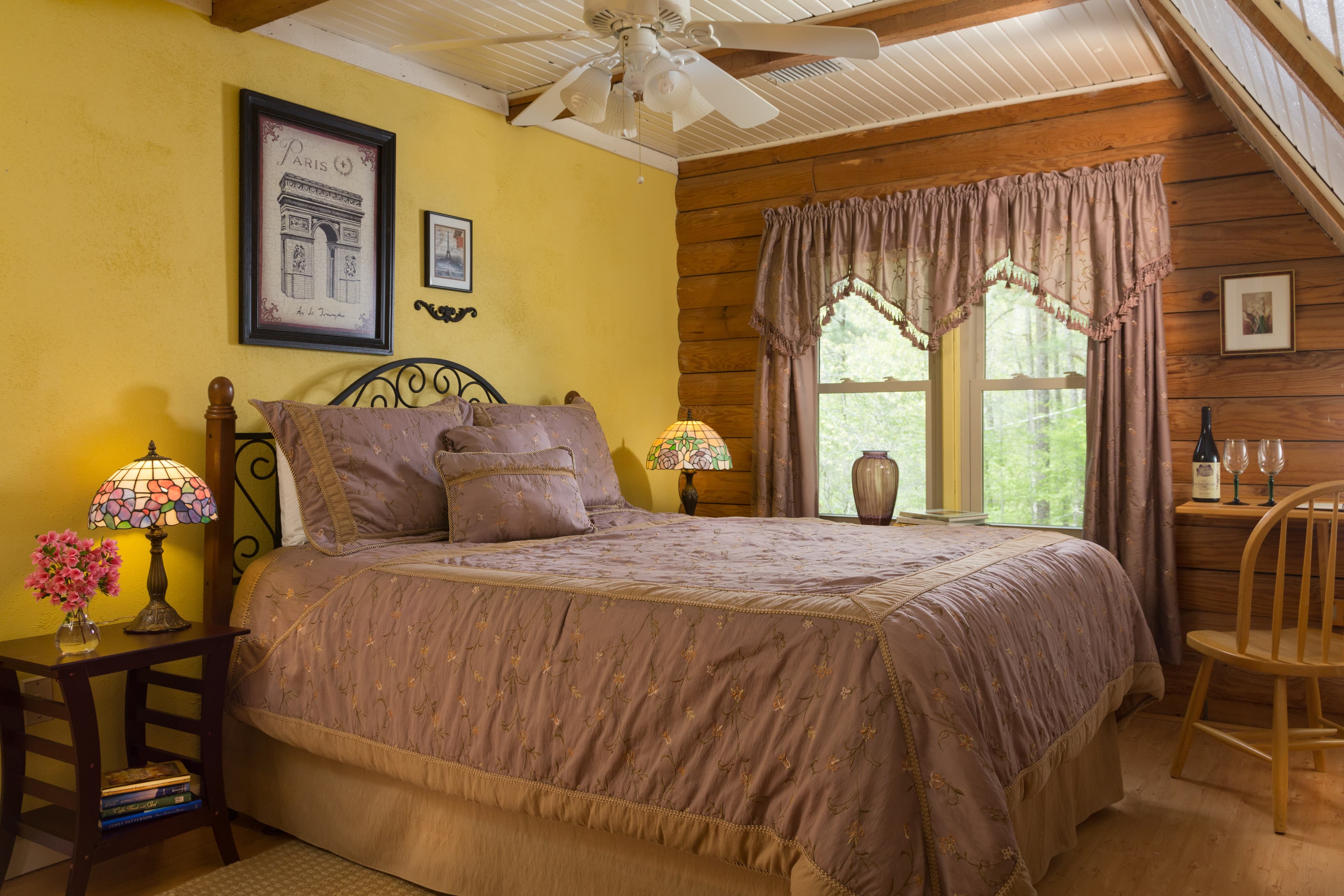 Bedroom with one yellow wall and one log wall with a window with purple curtains.  The queen-sized bed has a wrought iron headboard, and purple quilt and decorative pillows.  To the left of the bed is a wodden nightstand with a lamp and flowers on it.  There is another lamp to the right of the bed.  In the corner is a small wooden desk with a chair, and a bottle of wine & 2 glasses on top of the desk.