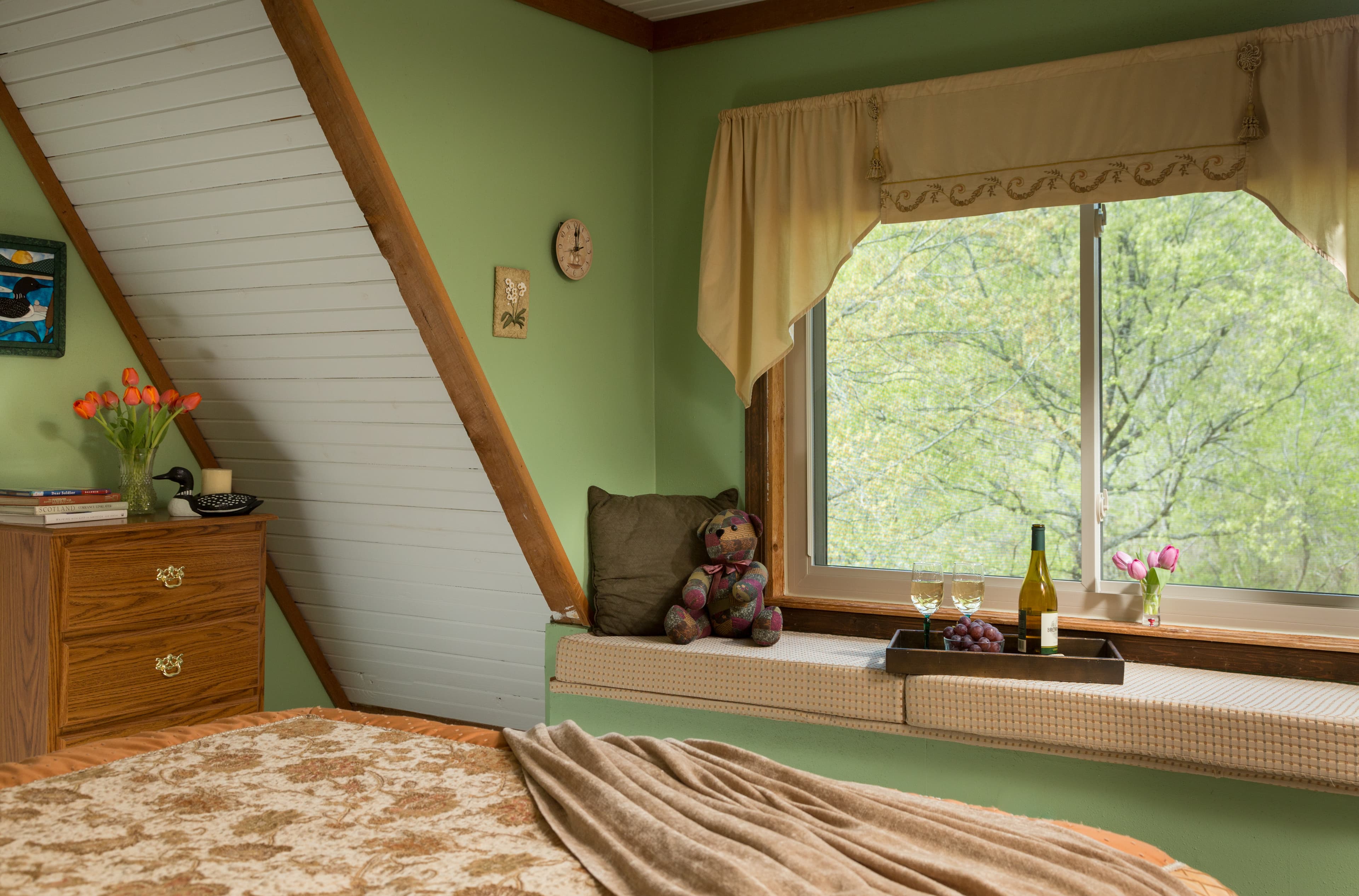 bedroom with part of a bed in the foreground.  On the bed are a quilt and throw blanket.  In front of the bed is a window seat with cushions.  On top of the cushions are a pillow, a stuffed bear, and a tray with a wine bottle and two glasses.  There is a large window above the window seat, overlooking trees.  To the left of the windowseat is a wooden dresser with flowers and books on top.