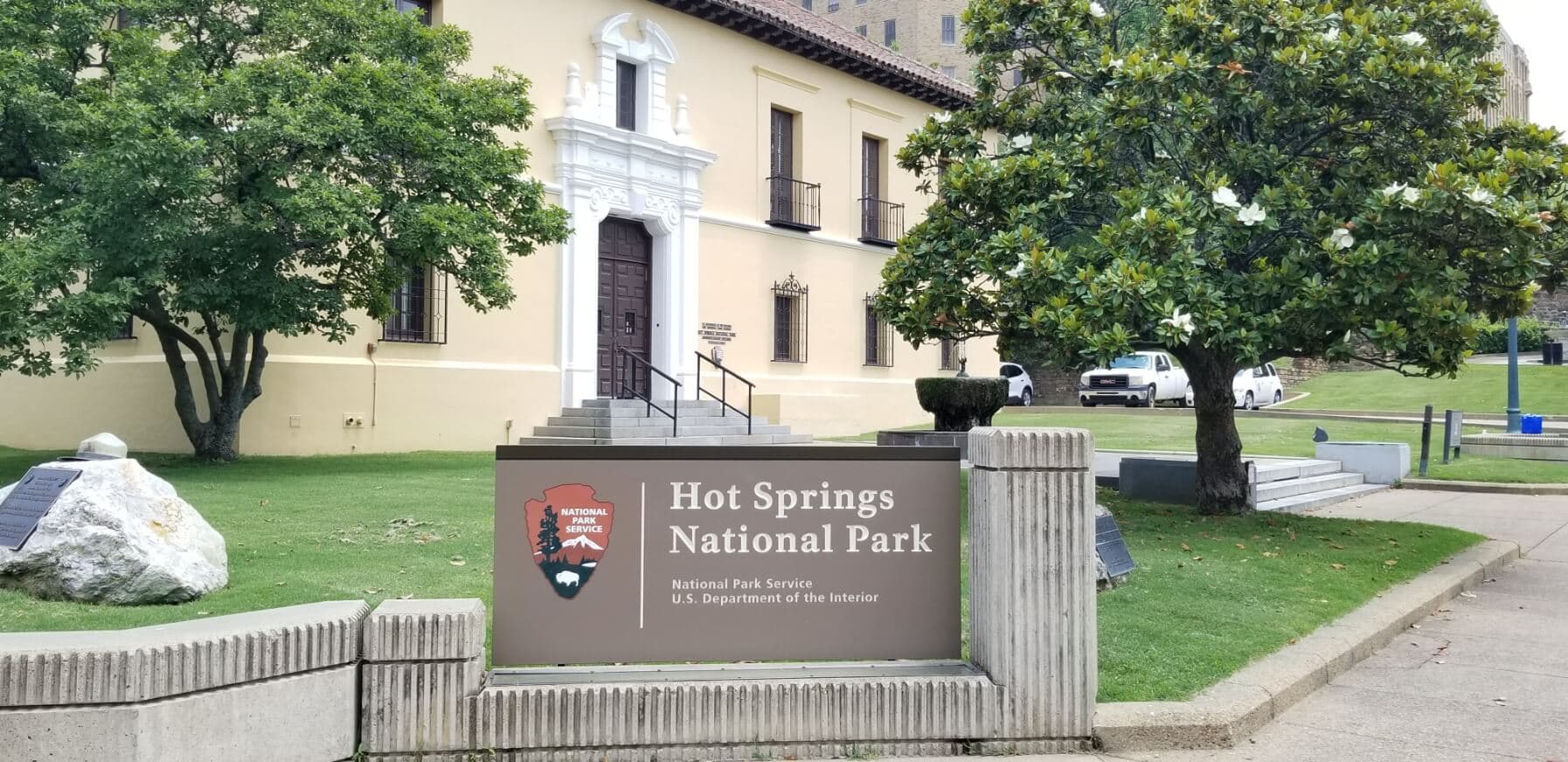 Sign for Hot Springs National Park in front of a historic building with trees and parking area. Sign for Hot Springs National Park in front of a historic building with trees and parking area.