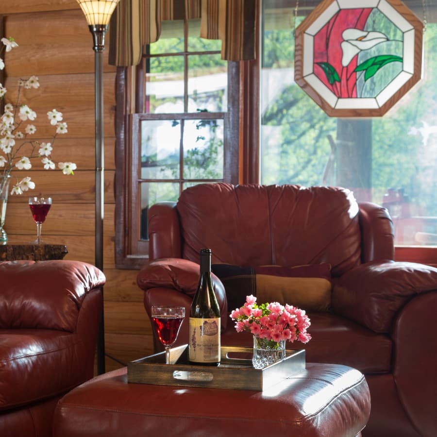 A cozy living room with leather furniture, stained glass in the window, and a tray with a bottle of wine, glasses of red wine, and some pink flowers in a crystal glass.