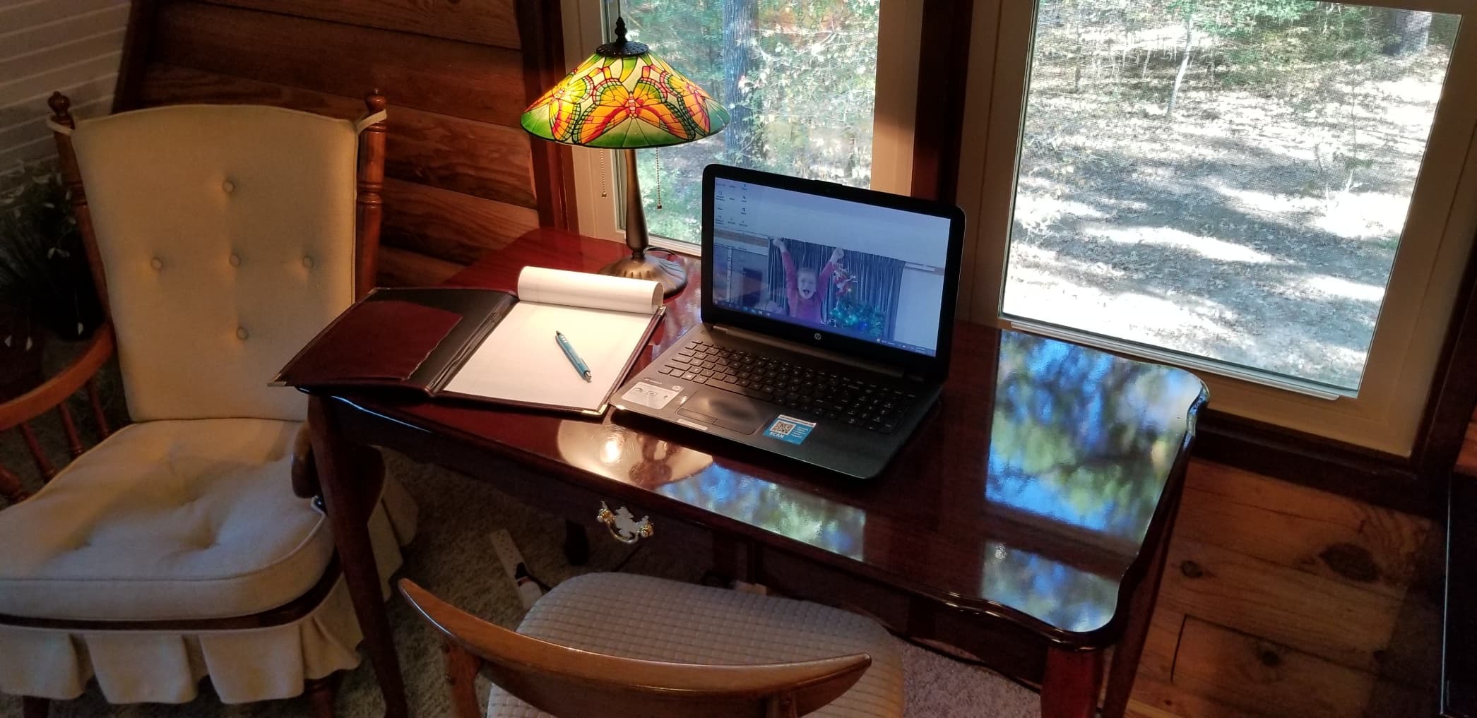 A cozy workspace featuring a wooden desk, laptop, notepad, and a stained glass lamp beside a window with a view of trees.