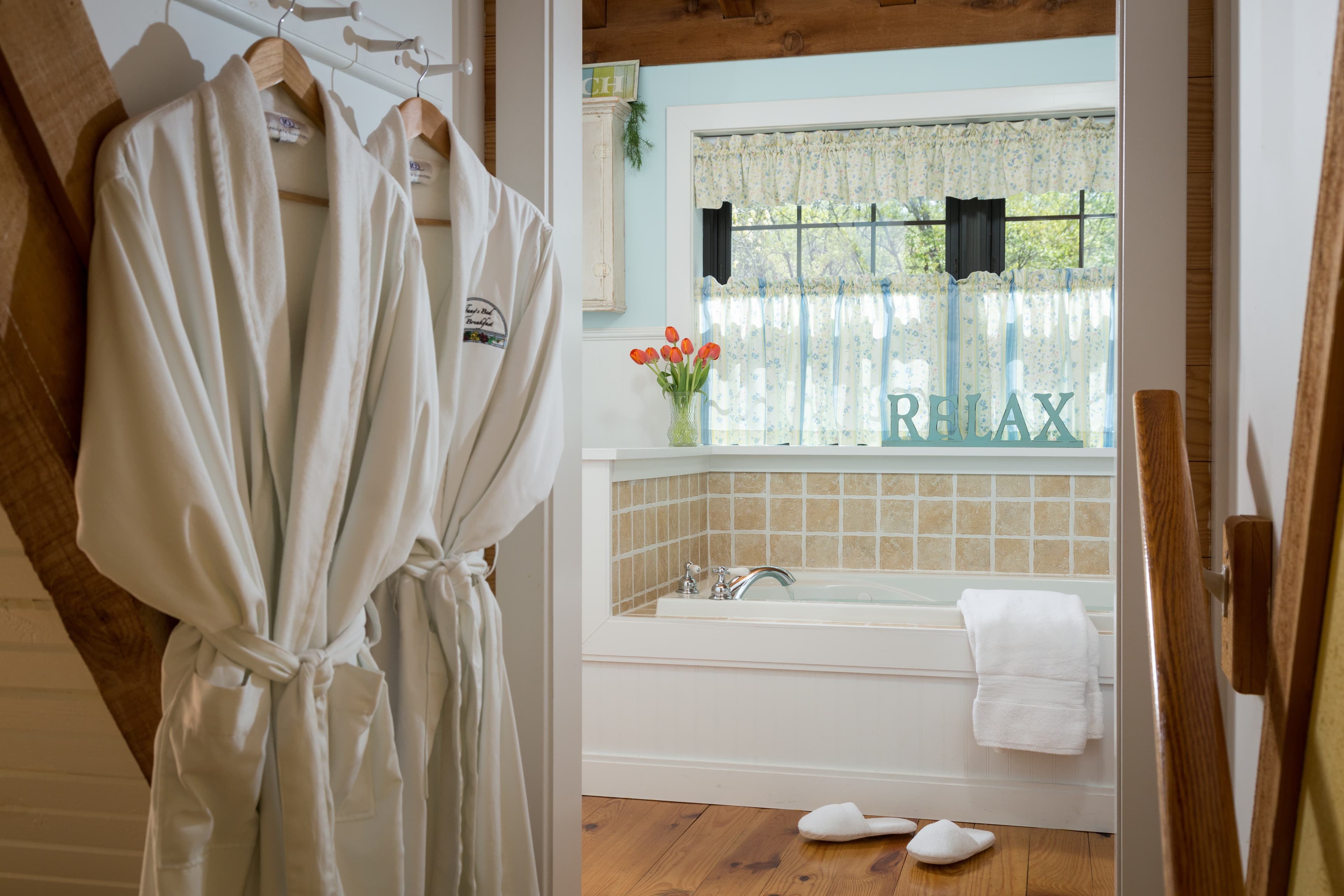 A cozy bathroom view featuring white robes, a hot tub, and a "RELAX" sign by a window with flowers.