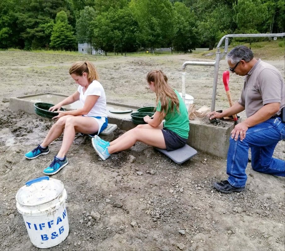 Three individuals are panning for gems at a muddy site surrounded by trees. Three individuals are panning for gems at a muddy site surrounded by trees.
