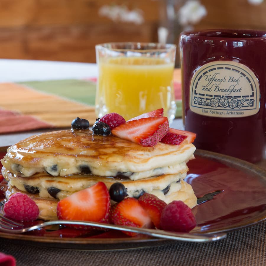 A plate of blueberry pancakes topped with sliced strawberries, with a glass of orange juice and a red coffee mug that says Tiffany's Bed & Breakfast.