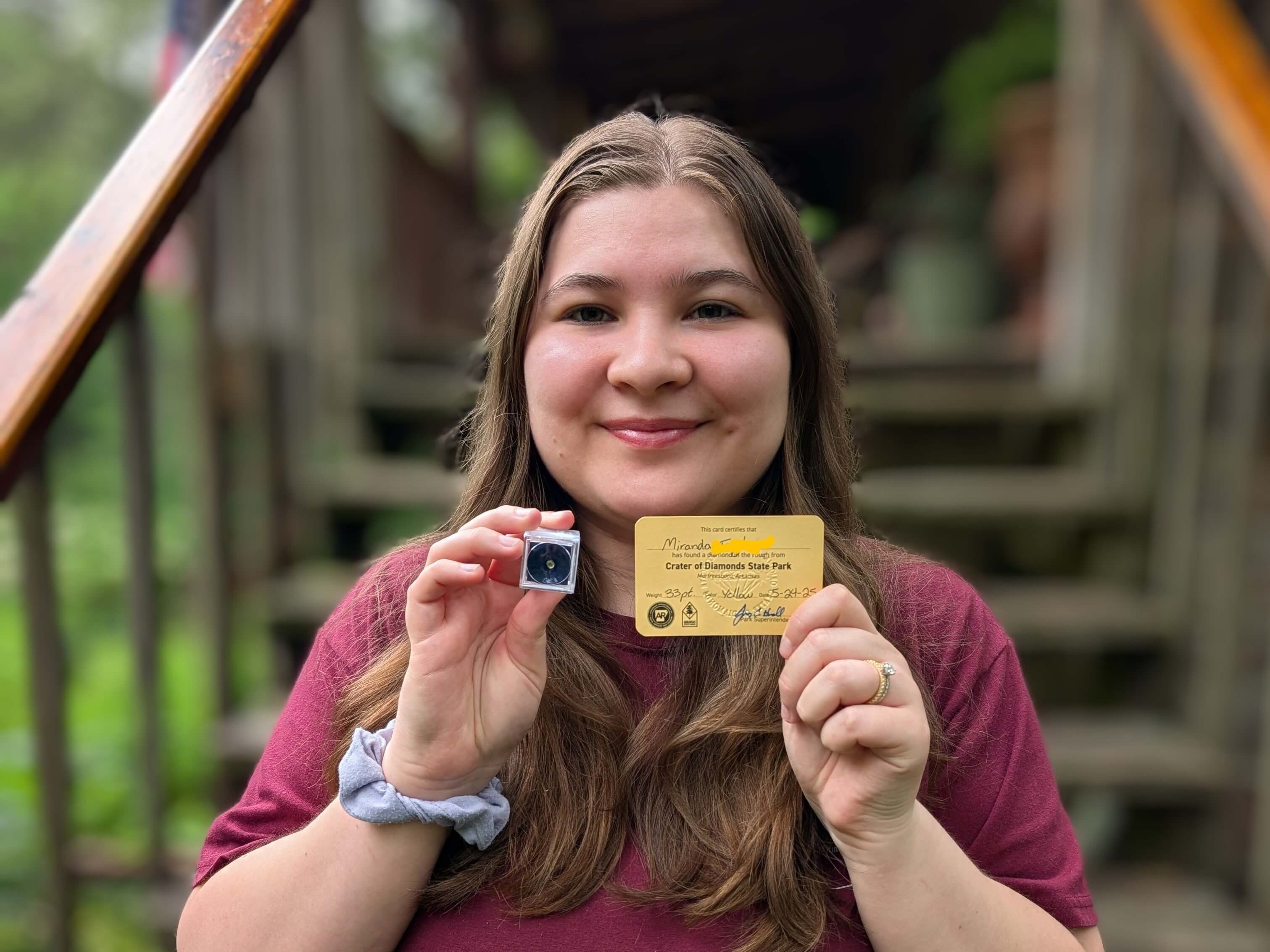 A young woman smiles while holding a small camera and a card in front of a wooden staircase.
