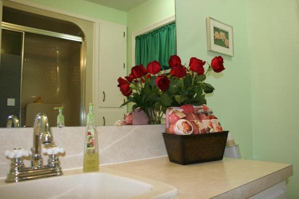 A bathroom with green walls and white trim. A shower is reflected in the vanity mirror. On the vanity is a sink, a basket of soaps, and a vase with red roses.