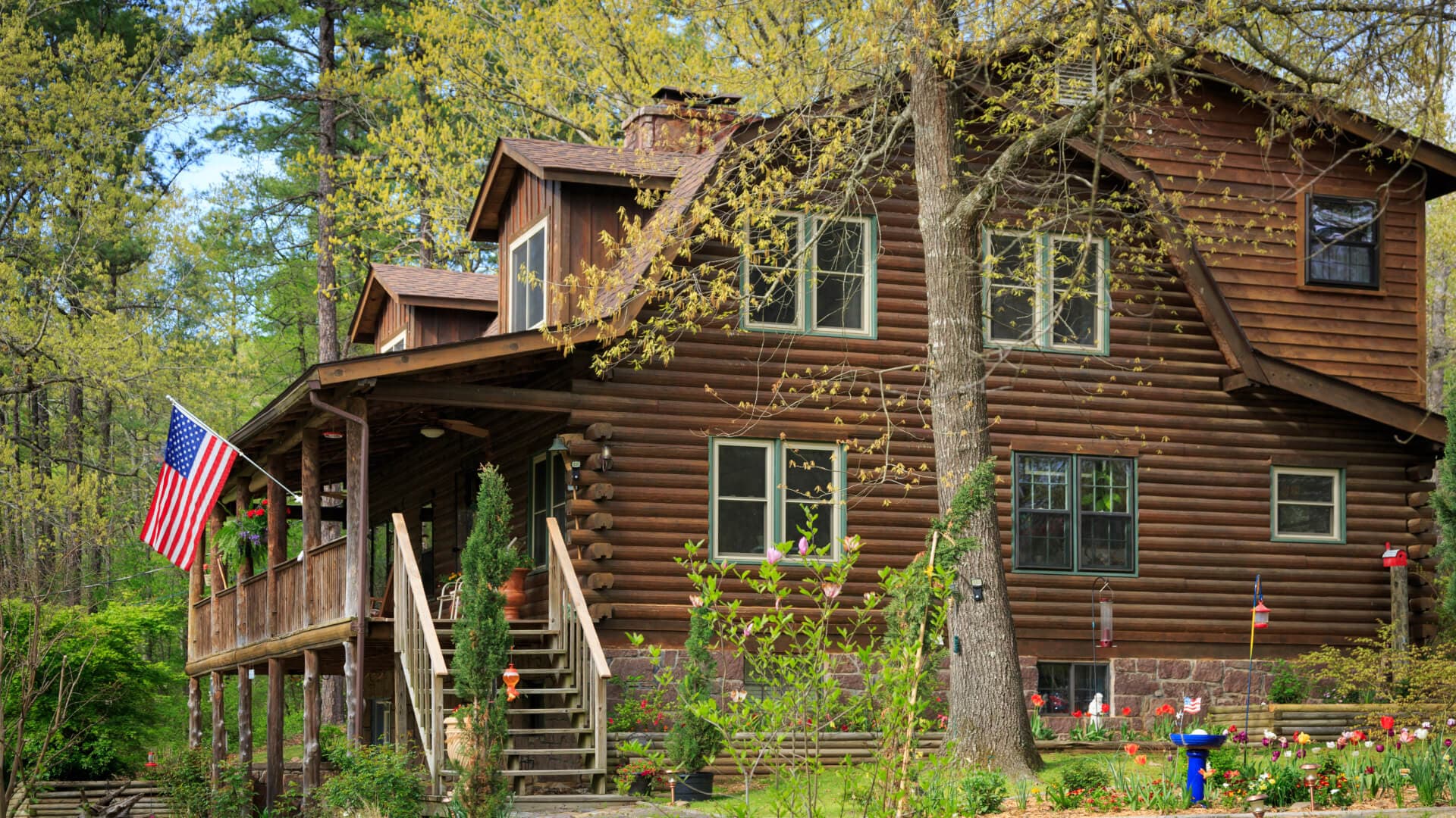 A multi story log house with a covered porch and American flag, surrounded by trees and gardens.