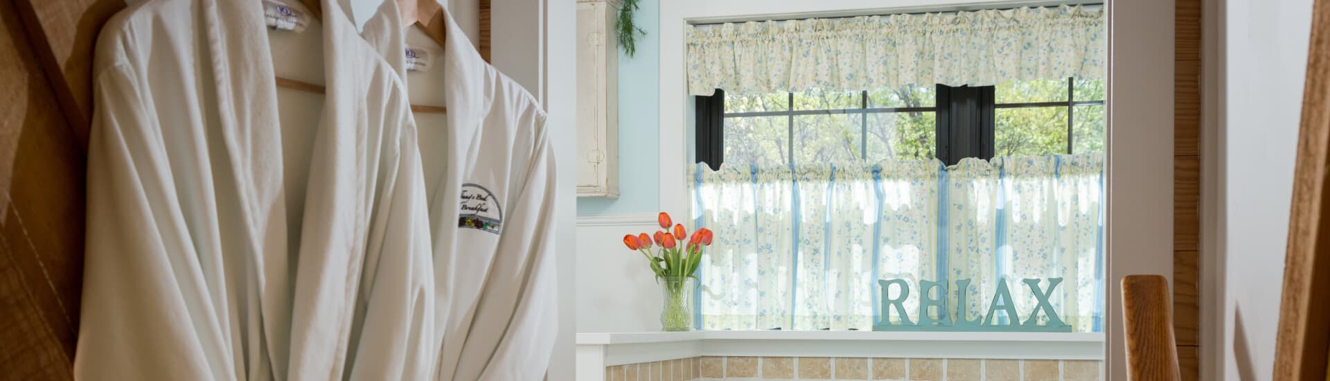 A bathroom with bathrobes hanging on hooks, a window with a shelf with orange tulips and a sign that says relax.