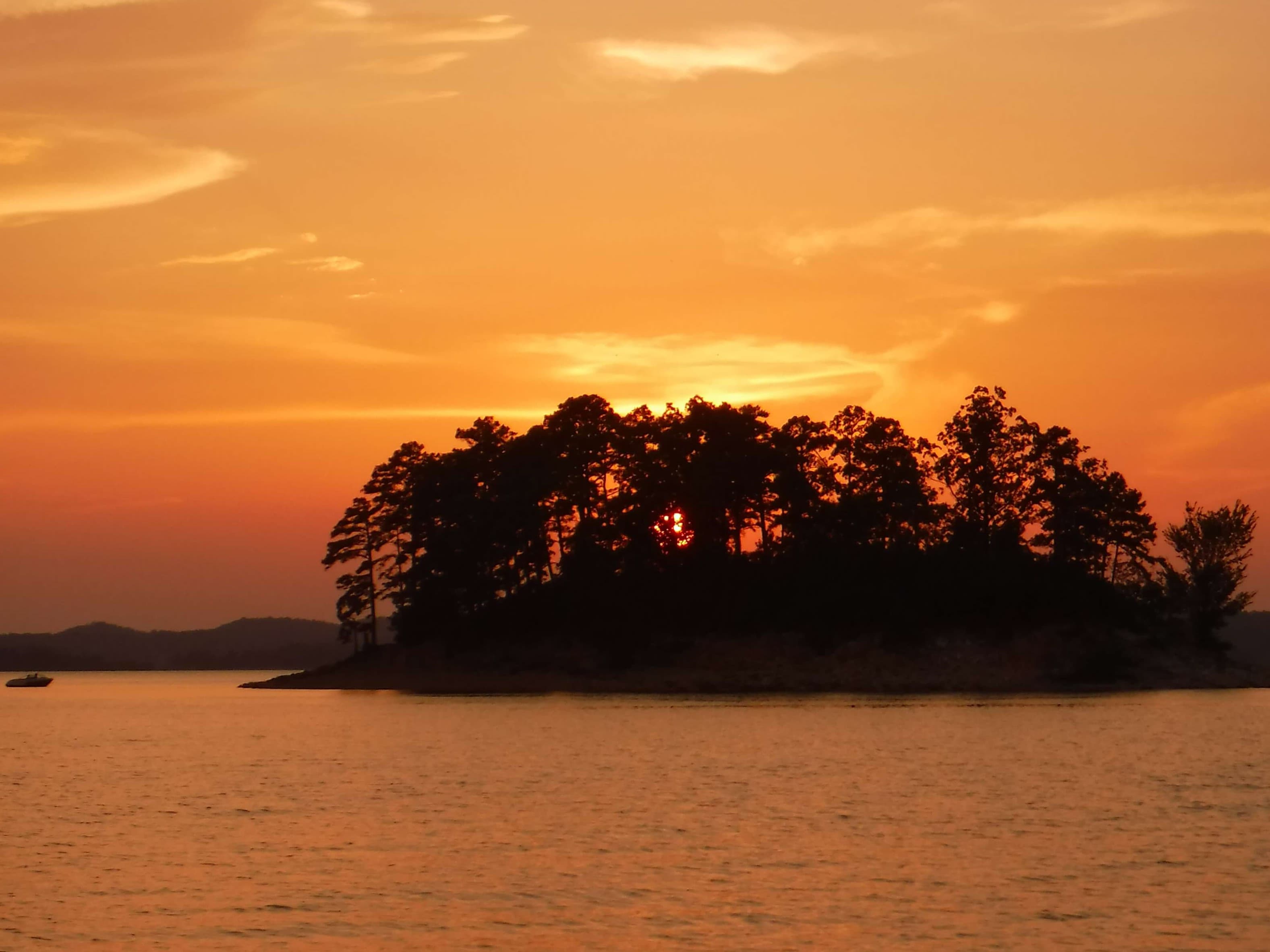 sunset behind a small island covered with trees.  A small boat sits in the water to the left of the island sunset behind a small island covered with trees.  A small boat sits in the water to the left of the island