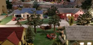 a miniature neighborhood on a model train set platform with a house, cars, lawnmower, and a person in a hammonck between 2 trees