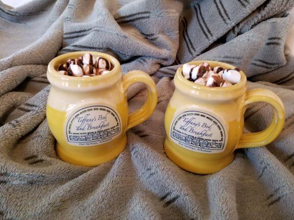 2 yellow coffee mugs filled with hot chocolate, topped with marshmallows and chocolate syrup, sitting on a green blanket