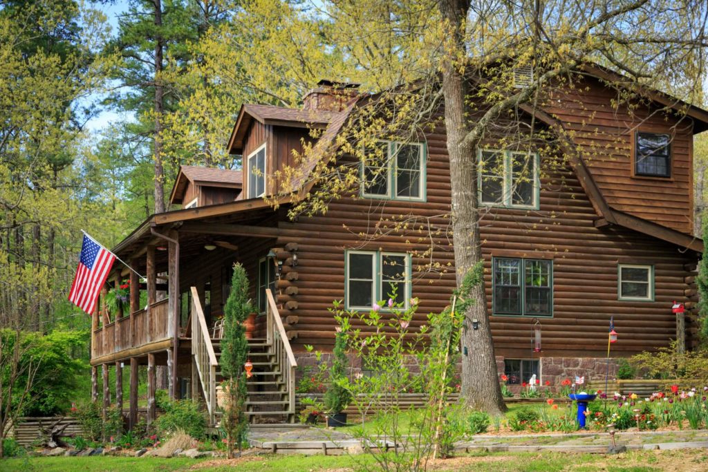 the side of a 2-story log home with trees and flowers in the foreground