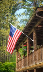 A US flag hanging from a wooden beam on the front porch of a log home. A hanging basket of red flowers is in the background