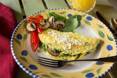 omelet with spinach, mushrooms and feta cheese on a yellow, blue & green plate. Mushrooms, spinach and red peppers garnish the plate and there is a fork beside the omelet