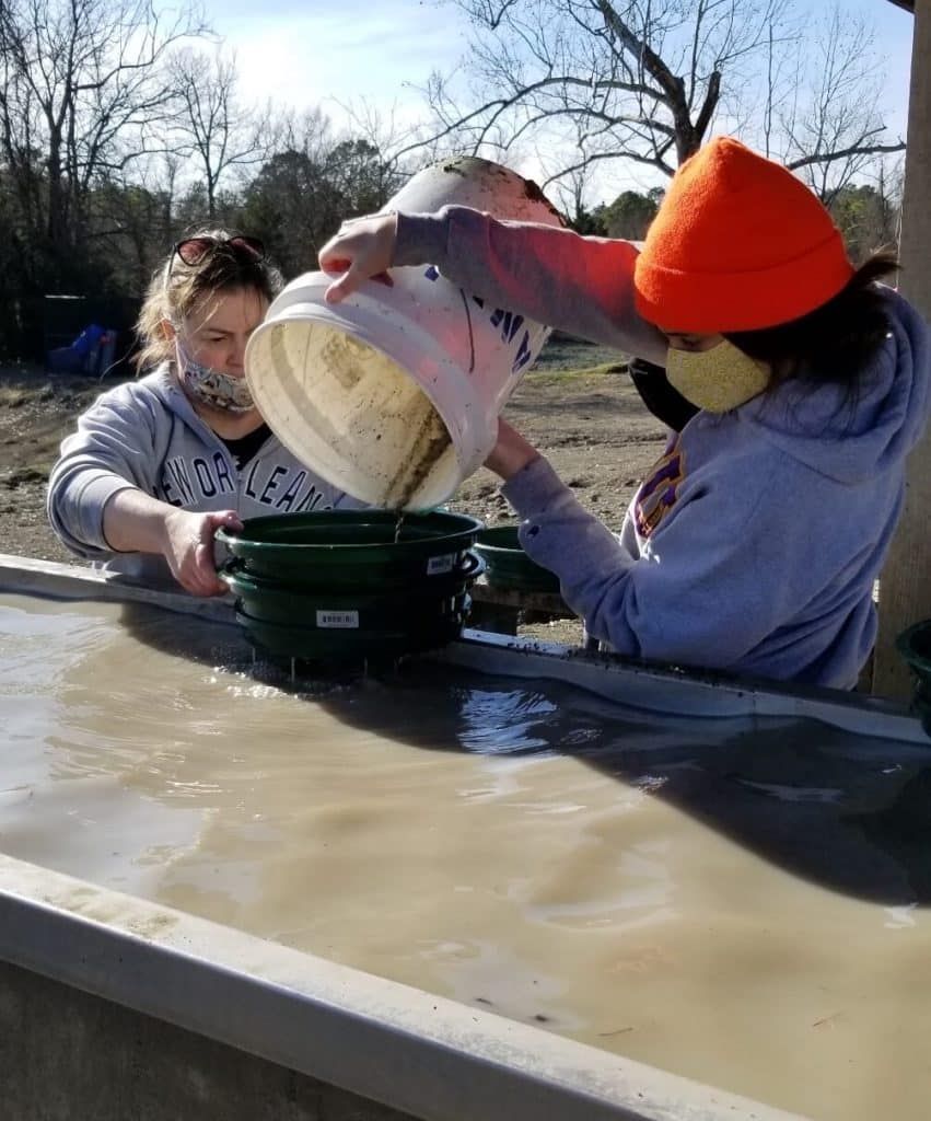 one person is holding 2 green screens above a trough of water while another person pours gravel out of a bucket into the screens