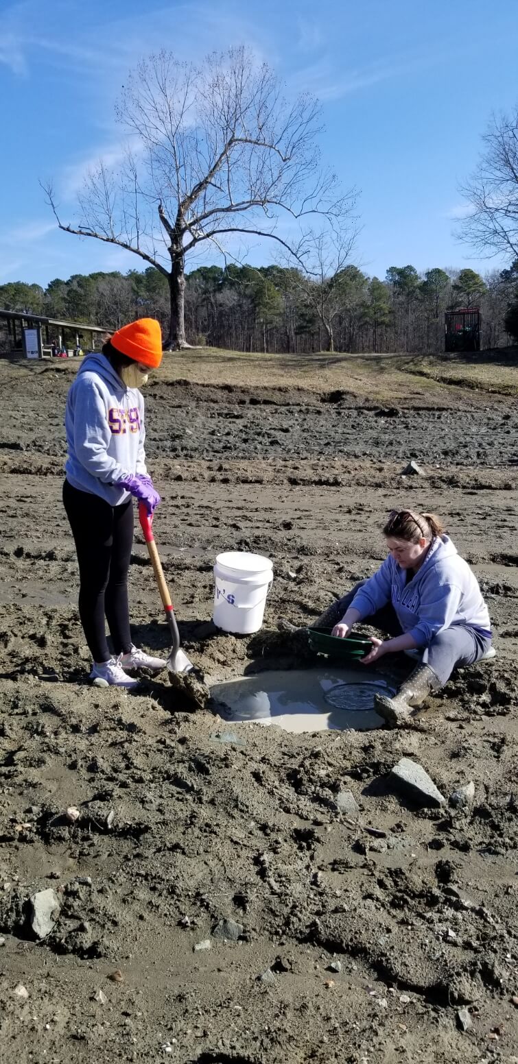 one person standing in a field holding a shovel, the other person is sitting on tne ground with a white bucket beside her, and a mudhole in front of her