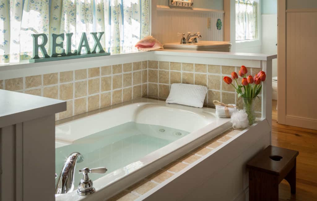 Jacuzzi tub below a window with a wooden sign that says RELAX. A vase of orange tullips sit on the side of the tub, along with washcloths and a bath pillow.