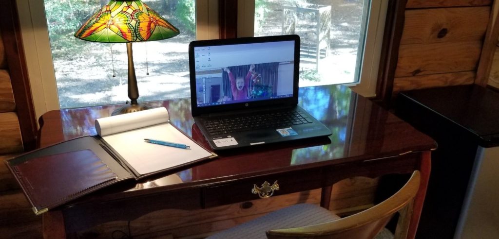 Laptop computer and tablet with pen on top of a writing desk, facing 2 windows looking out into woods